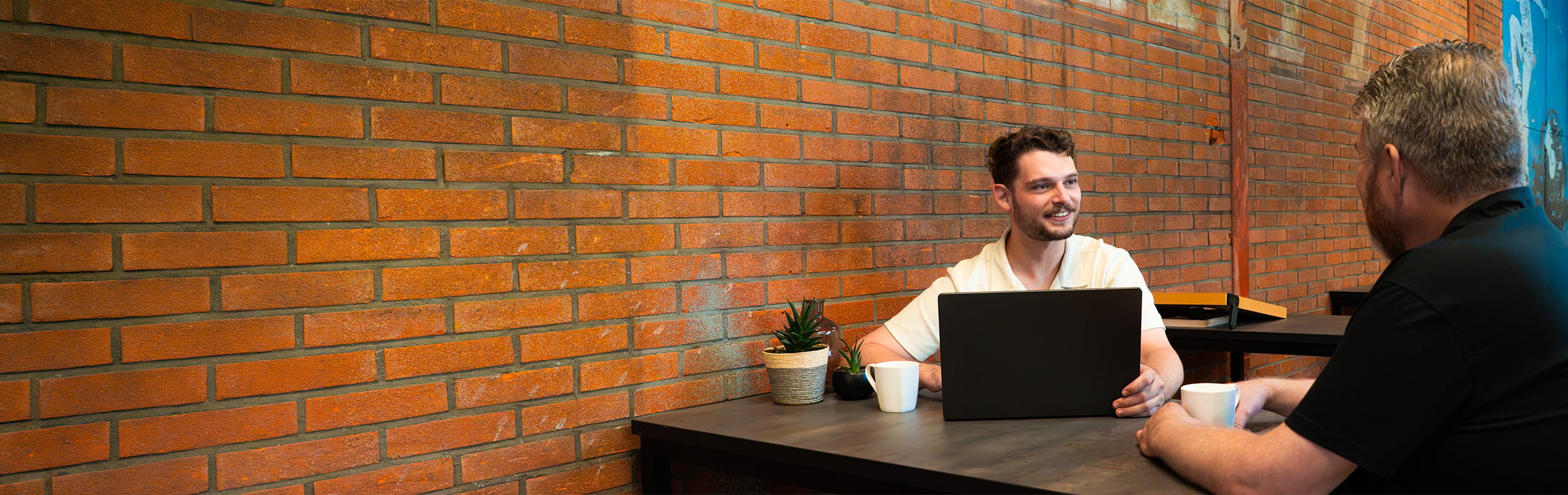 Persoonlijk gesprek tussen een Azerty-accountmanager en een klant, aan tafel met laptop en koffie voor een bakstenen muur